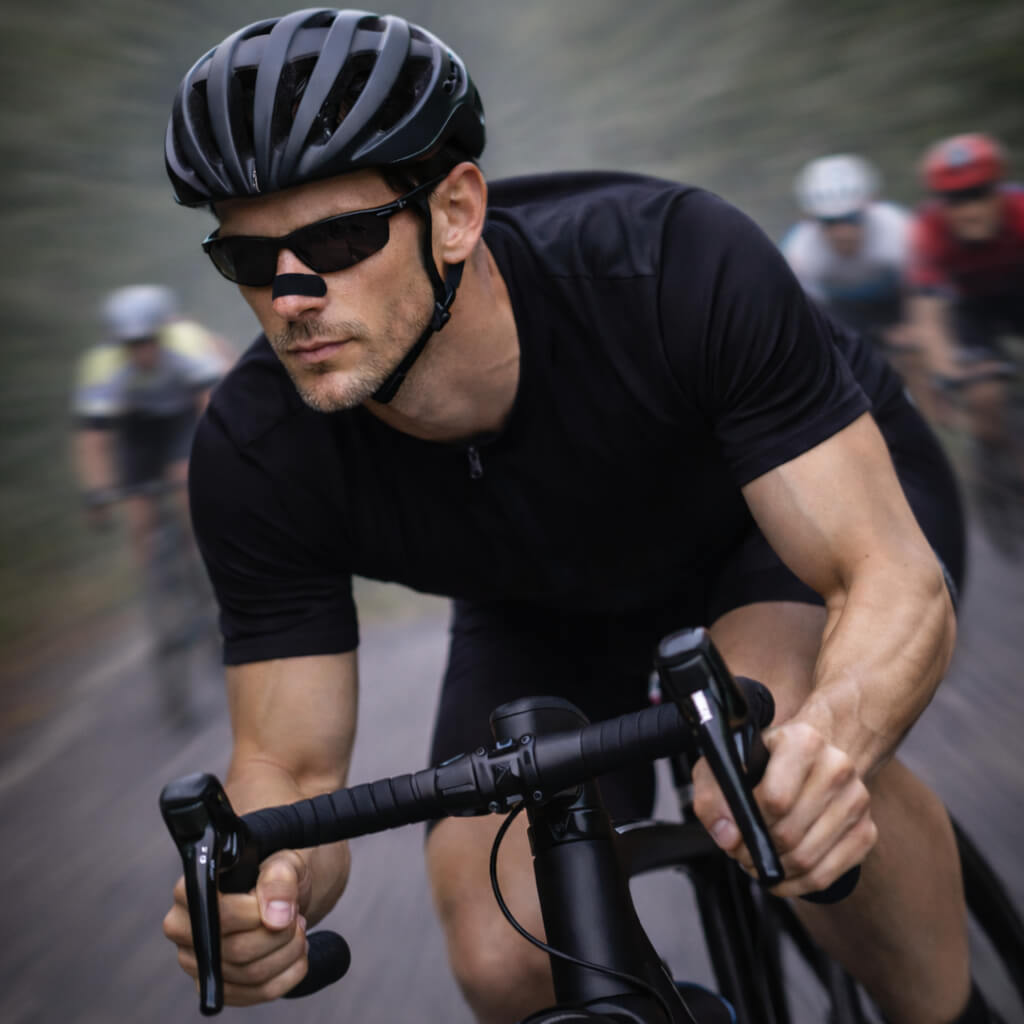 A male cyclist is powering through a road race while wearing a black BREATHMODE nasal strip