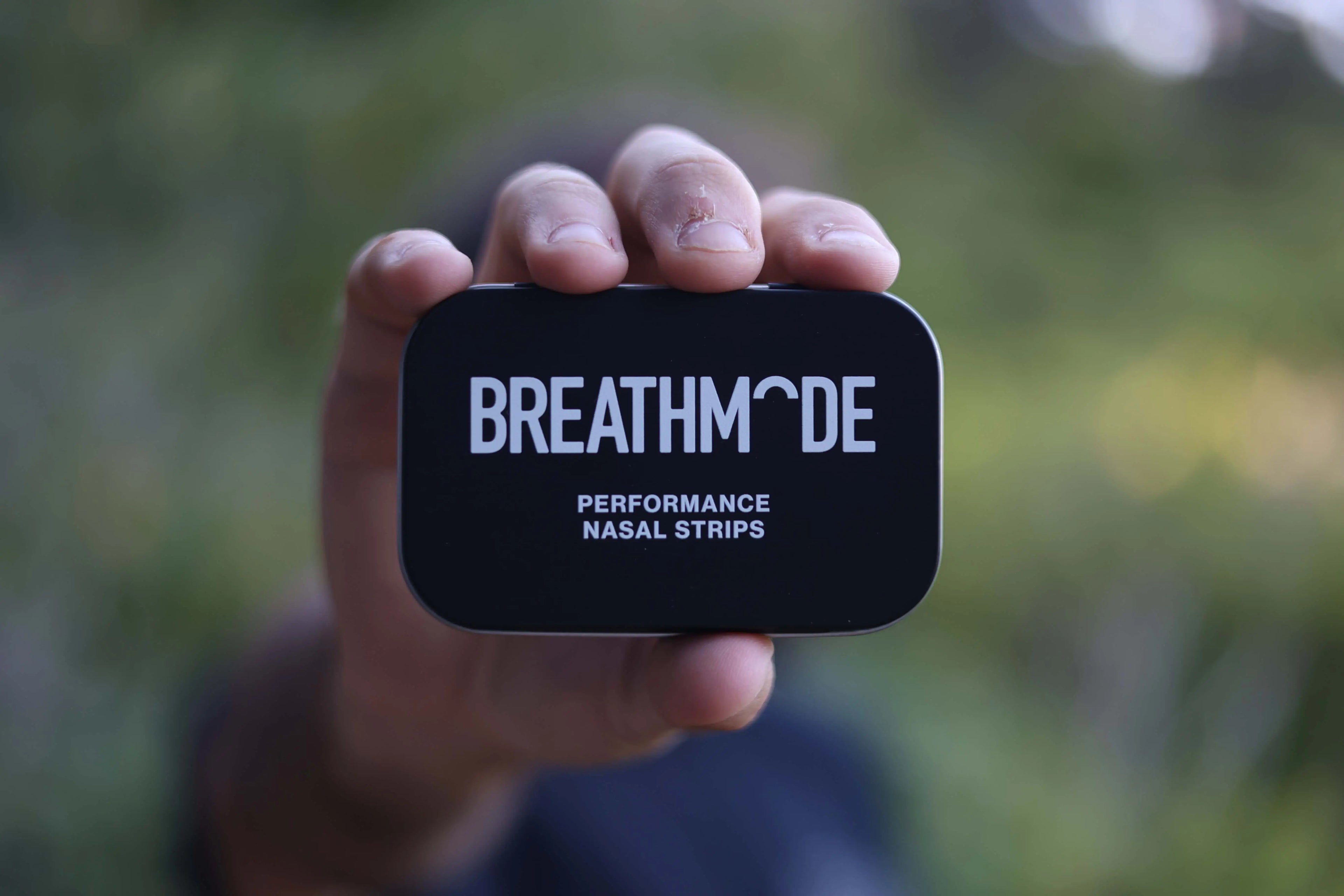 BREATHMODE Founder Jozef Wright's hand holding a black nasal strip tin with 'BREATHMODE' branding against a blurred green background